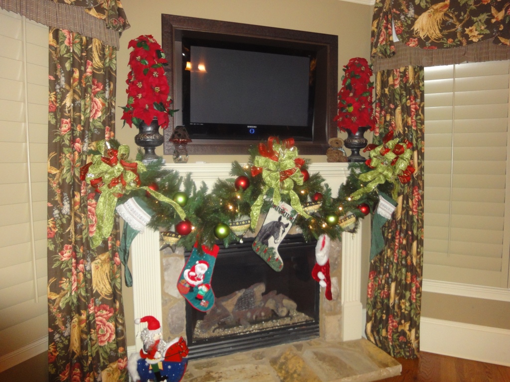 Mantle hosting a tv over top, framed by picture molding, decorated with traditional stockings, and red/green ribbon.
